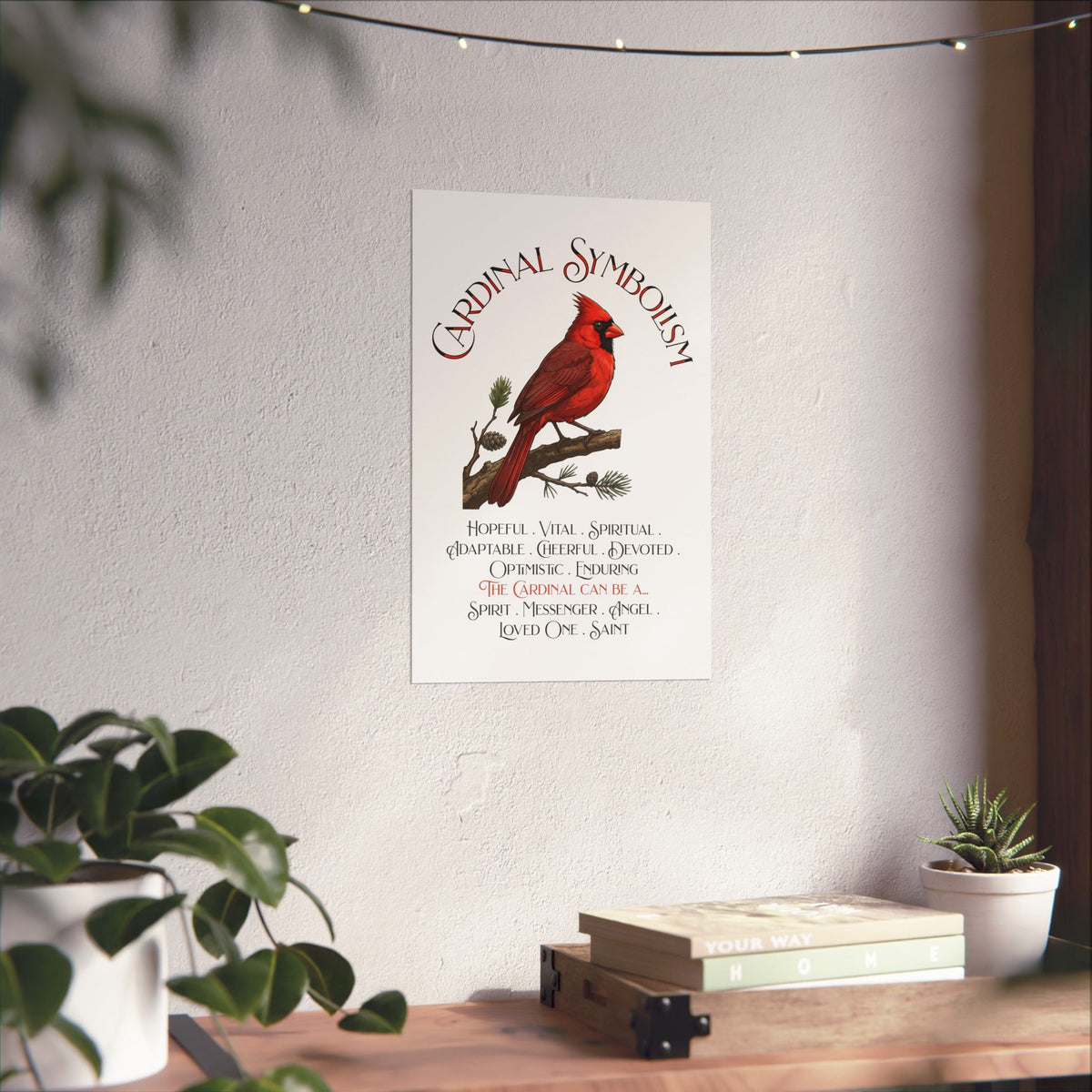 A vertical wall art print featuring a red cardinal on a pine branch with the title 'Cardinal Symbolism' and a list of cardinal qualities and meanings, displayed on a light wall above a wooden shelf with books and plants.