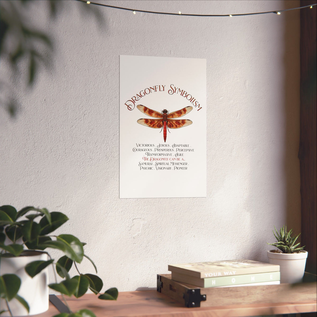 A vertical wall art print featuring a red dragonfly with the title 'Dragonfly Symbolism' and explanatory text below, displayed on a textured light wall above a wooden surface with books and plants.