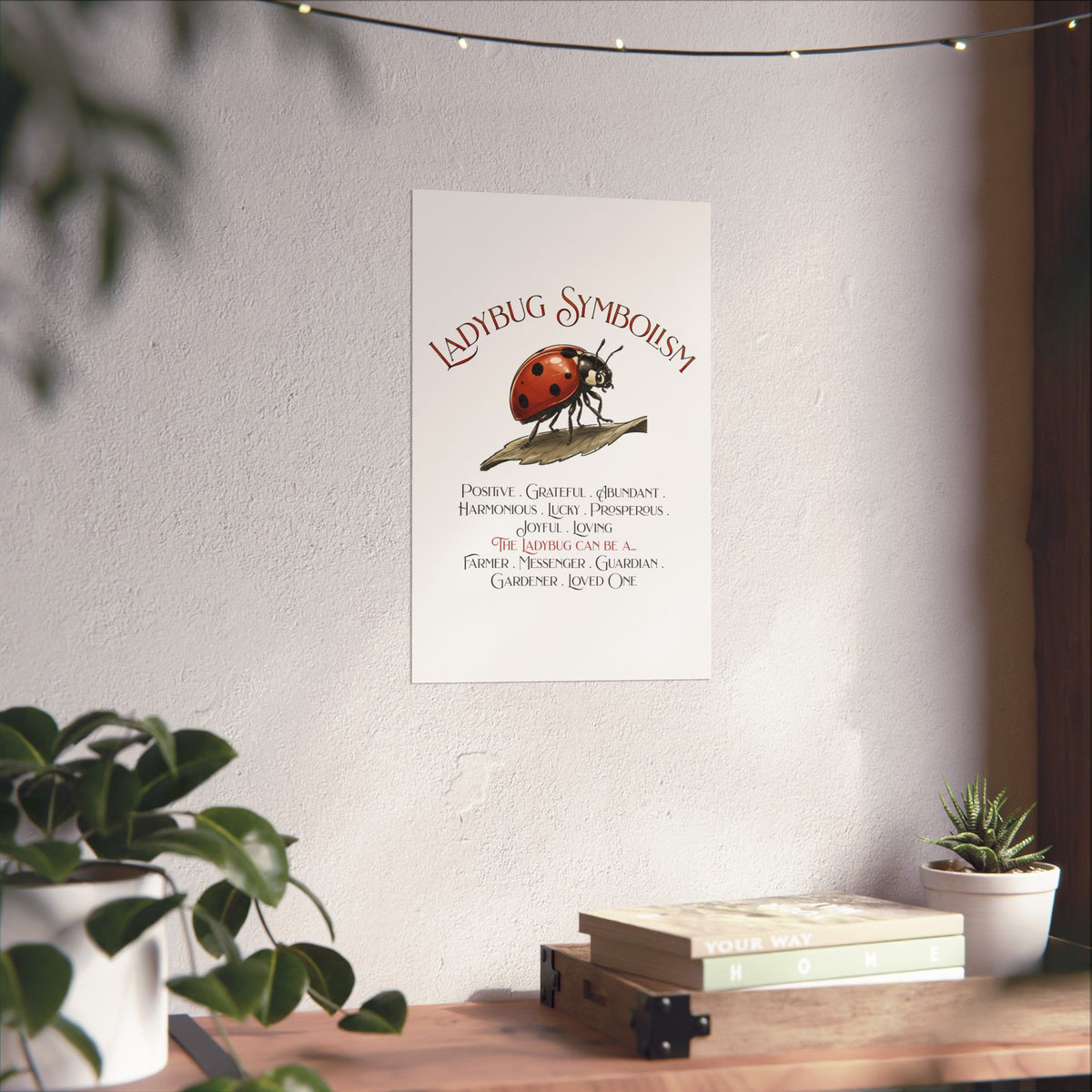 A vertical wall art print featuring the Ladybug Symbolism text and an illustration of a red ladybug on a leaf, displayed on a textured light wall above a wooden surface with books and plants.