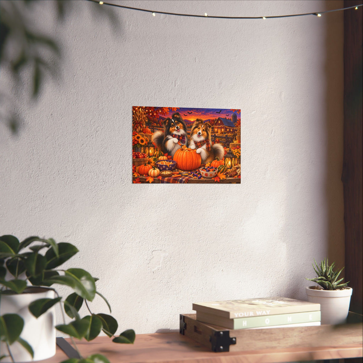 A rectangular wall art print showing two shelties carving a jack o' lantern, displayed on a white wall above a wooden surface with books and small plants.