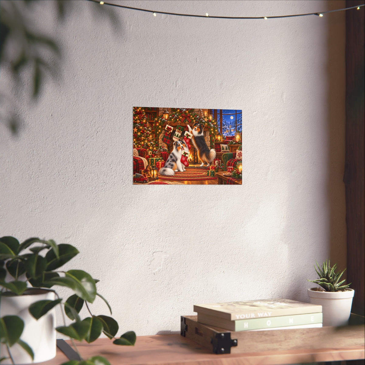 A rectangular wall art print showing a festive Christmas scene with shelties hanging stockings, displayed on a textured off-white wall above a wooden table with books and small potted plants.