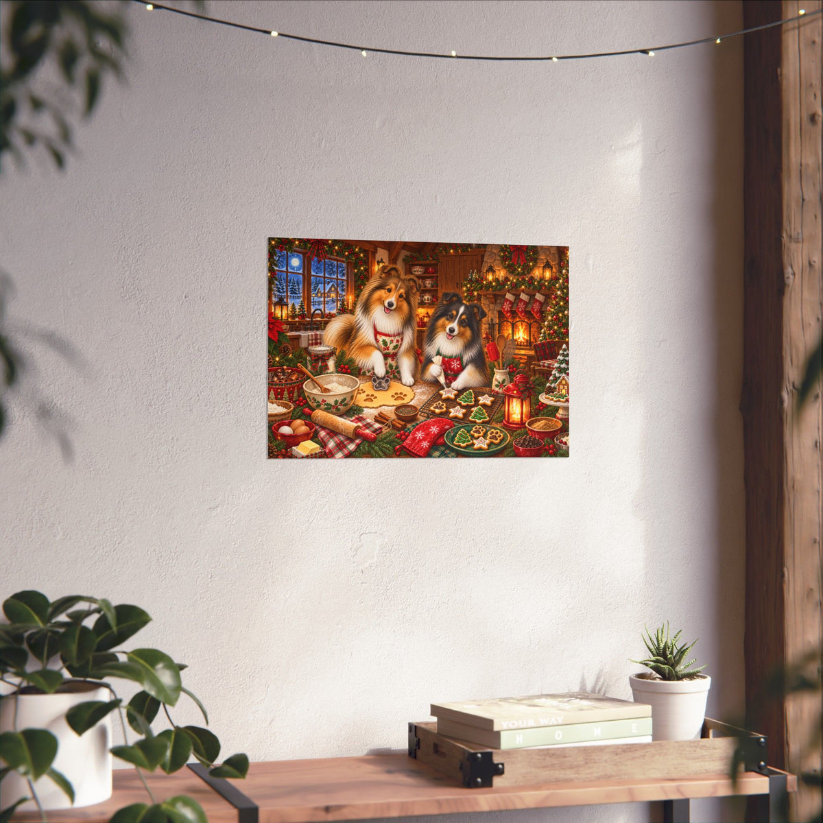 A rectangular wall art print showing two shelties baking and decorating cookies at a festive holiday table, displayed on a textured white wall above a wooden surface with books, a small plant, and hanging string lights.