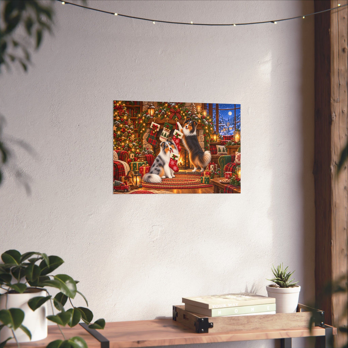 A rectangular wall art print showing a festive Christmas scene with shelties hanging stockings, displayed on a textured off-white wall above a wooden table with books and small potted plants.