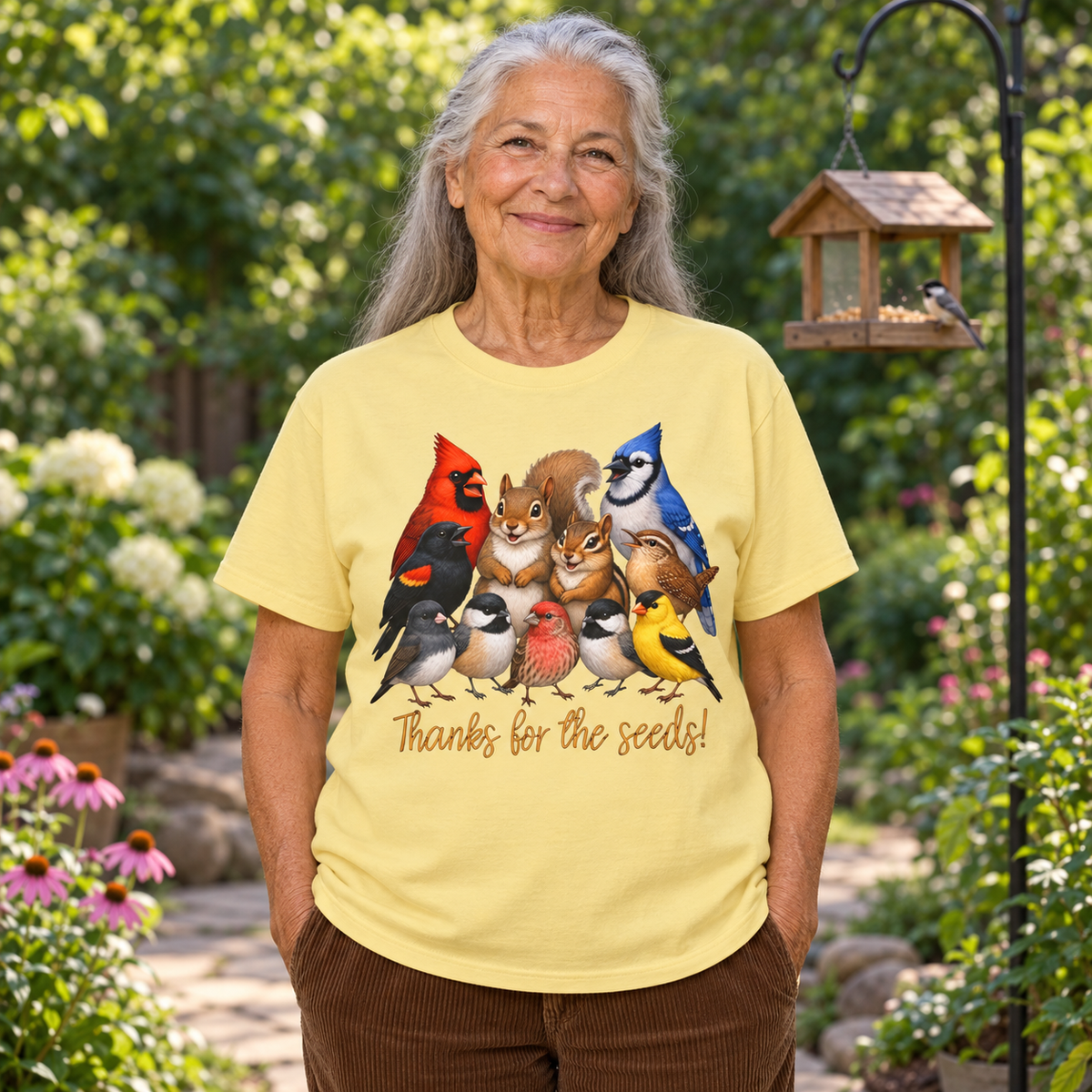 An older woman stands in a garden wearing a light yellow graphic T-shirt with a colorful illustration of various birds, a squirrel, and a chipmunk with text below that says, 'Thanks for the seeds!'.