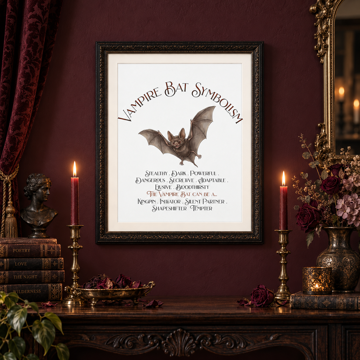 A framed gothic art print titled 'Vampire Bat Symbolism' featuring a bat illustration and text, displayed on a dark wooden console table against a deep burgundy wall, flanked by lit candles, books, and floral arrangements.