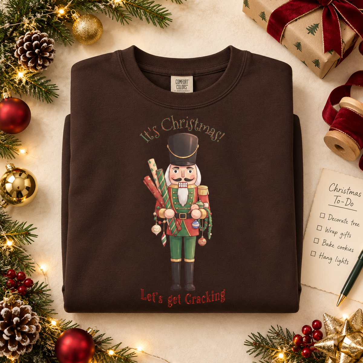 A folded dark brown crewneck sweatshirt with a Christmas nutcracker holding decorations and wrapping paper surrounded by the text 'It's Christmas! Let's get Cracking'. The pullover is styled in a festive flat-lay with pine branches, ornaments, wrapped gifts, and a 'Christmas To-Do' list.