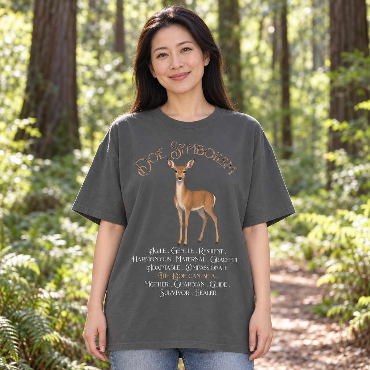 A woman standing in a sunlit forest wearing an oversized dark charcoal gray t-shirt with a graphic of a deer and the text 'Doe Symbolism' and a list of qualities. She is also wearing blue jeans.
