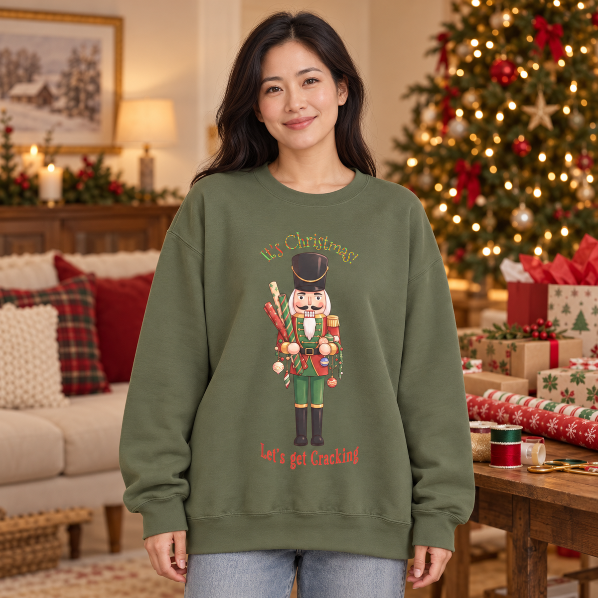 A woman wearing an olive green crewneck sweatshirt with a Christmas nutcracker holding gift wrap and decorations surrounded by the text 'Let's Christmas! Let's get Crackled'. The woman stands in a cozy, decorated living room with a Christmas tree, ribbons, and gift wrap and bags in the background.