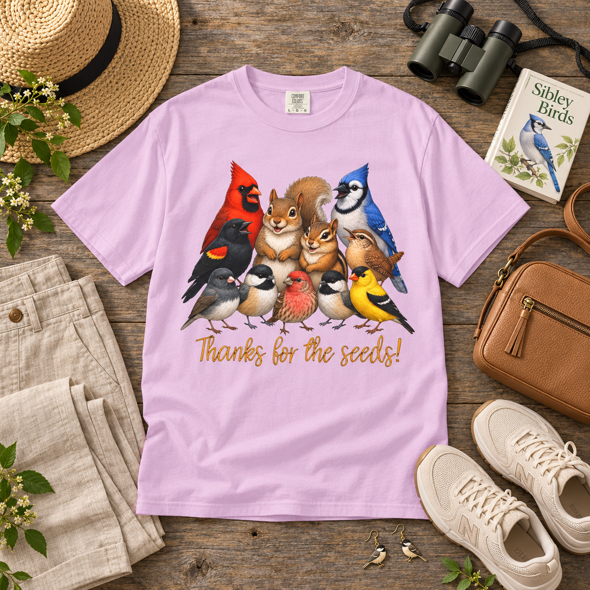 A light purple crewneck t-shirt laid flat on a rustic wooden table, featuring a colorful graphic of various birds, a squirrel, and a chipmunk with the text 'Thanks for the seeds!'. The shirt is styled in a flat-lay arrangement with a straw hat, beige pants, binoculars, a book, a brown leather purse, and white sneakers.