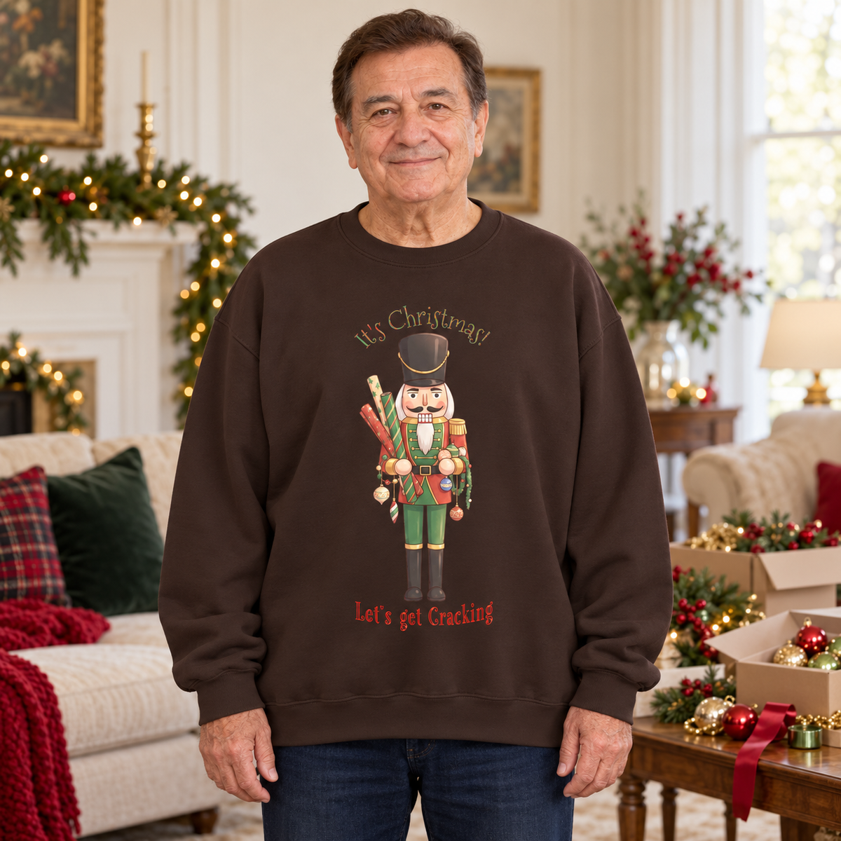 A man wearing a dark brown crewneck sweatshirt with a Christmas nutcracker graphic and the text 'It's Christmas Let's get Cracking' in a decorated living room setting.