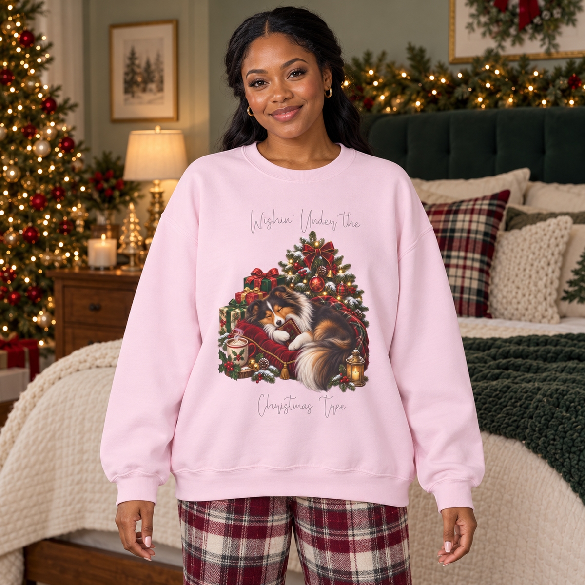 A woman wearing a light pink crewneck sweatshirt with a Christmas-themed graphic of a Shetland sheepdog sleeping under a Christmas tree and the text, 'Wishin' Under the Christmas Tree'. The woman is wearing red and white plaid pajama pants and is posed in a cozy, decorated bedroom with a Christmas tree and bed in the background.