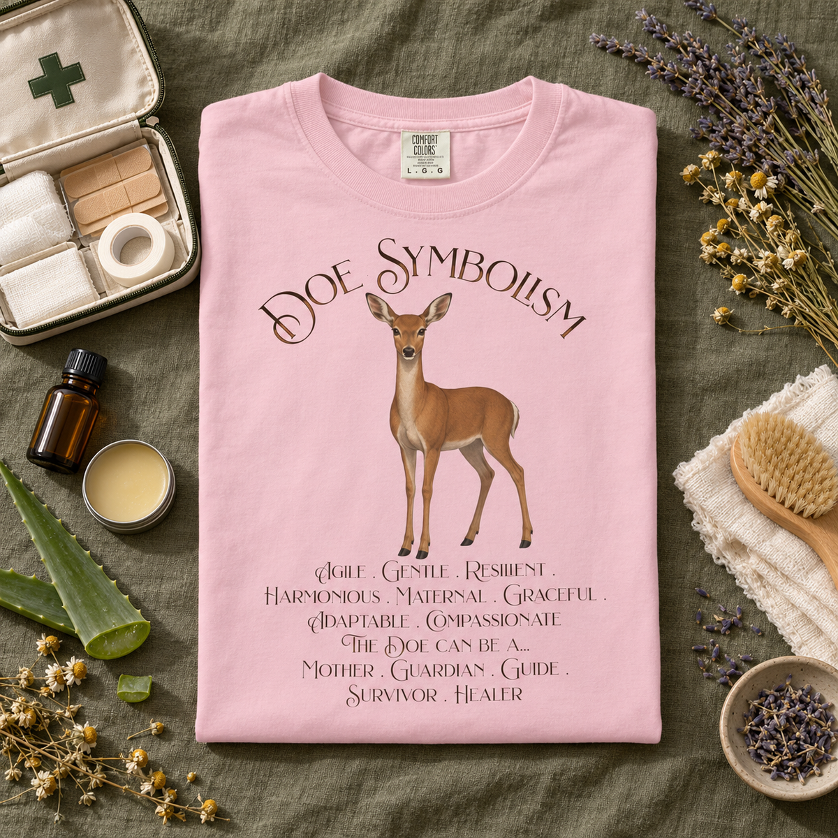 A folded light pink T-shirt with a deer illustration and the words 'Doe Symbolism' and a list of qualities printed on the front, styled in a flat lay on a green fabric background with dried flowers, a first-aid kit, a small bottle, and a brush.