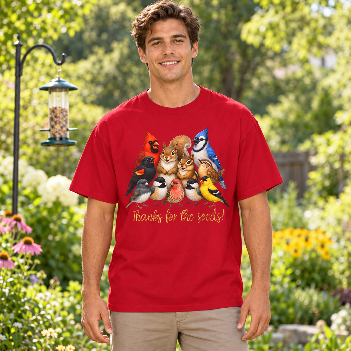 A man wearing a red crewneck t-shirt with a colorful graphic of various birds, a squirrel, and a chipmunk and yellow text 'Thanks for the seeds!' The man stands in a sunny garden.