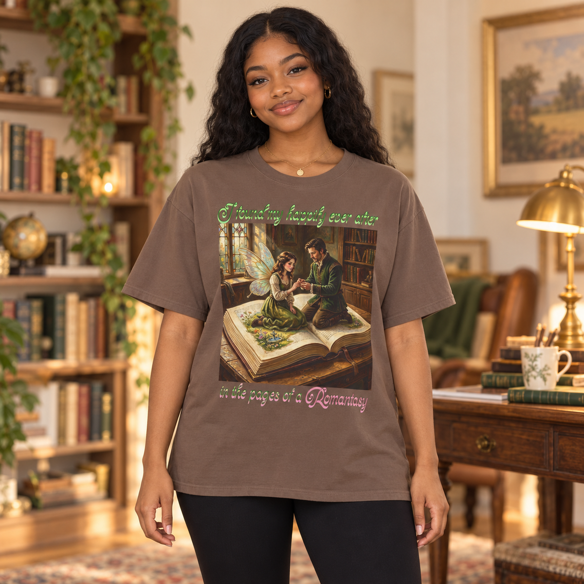 A woman stands in a cozy, book-filled room wearing a brown graphic t-shirt. The t-shirt features a vintage-style illustration of a man proposing to a fairy as they sit on an open book, with green and pink text reading 'I found my happily ever after in the pages of a Romantasy'.