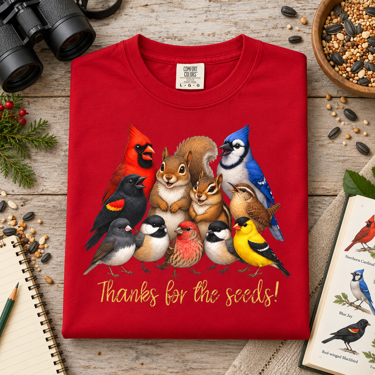 A folded red crewneck sweatshirt with a colorful graphic of various birds, a squirrel, and a chipmunk and yellow text saying, 'Thanks for the seeds!' The shirt is displayed on a rustic wooden table surrounded by a bowl of seeds, a bird book, binoculars, and a notebook.