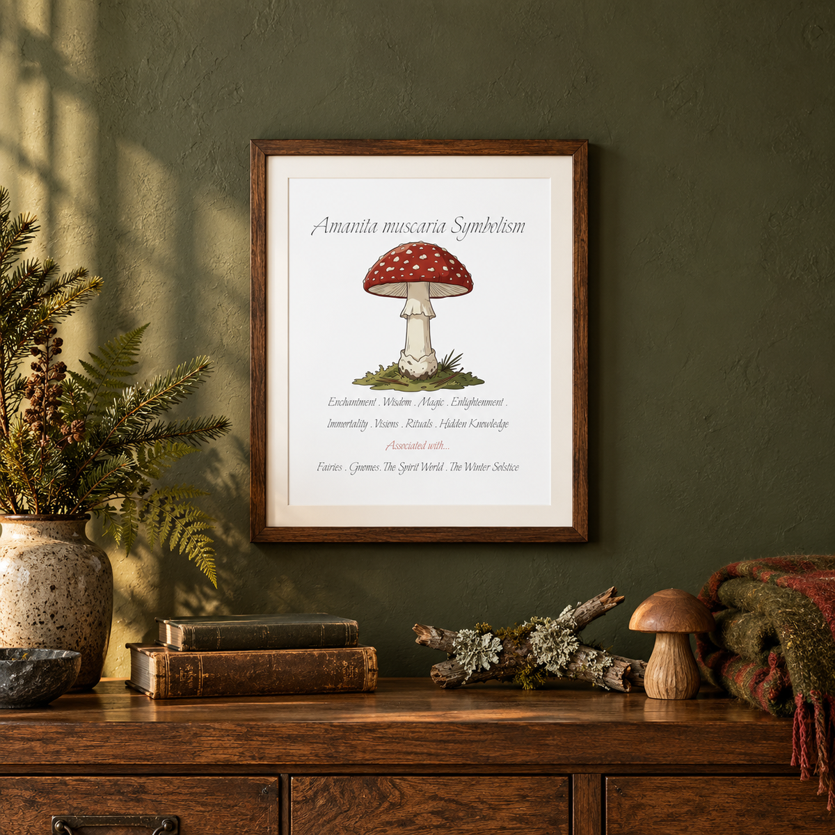 A framed botanical art print of Amanita muscaria symbolism on a dark olive green wall above a wooden console table with a vase of greenery, a mossy branch, a blanket, plants, and books.