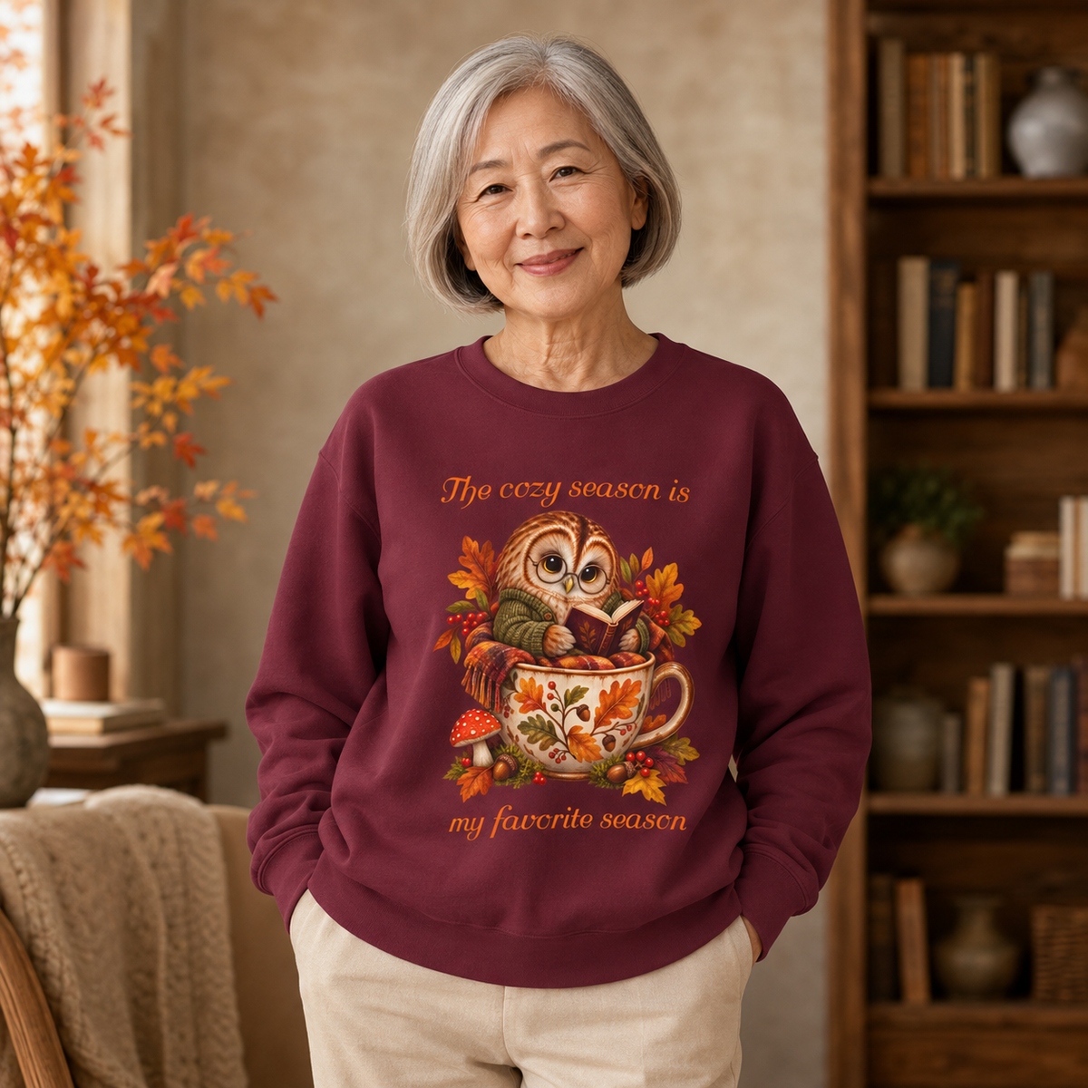 An older woman with short gray hair stands indoors wearing a burgundy crewneck sweatshirt with a colorful autumn owl graphic and the text, 'The cozy season is my favorite season.' The woman is also wearing light beige pants, with a warm, cozy room background featuring a bookshelf and autumn leaves.
