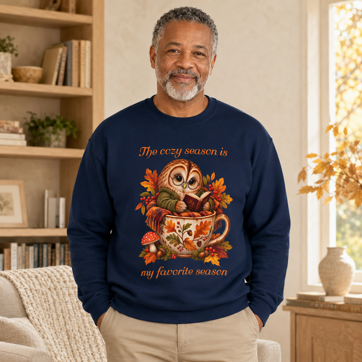 A man wearing a navy blue crewneck sweatshirt with a colorful graphic of an owl in a teacup surrounded by autumn leaves, with the text, 'The cozy season is my favorite season'.
