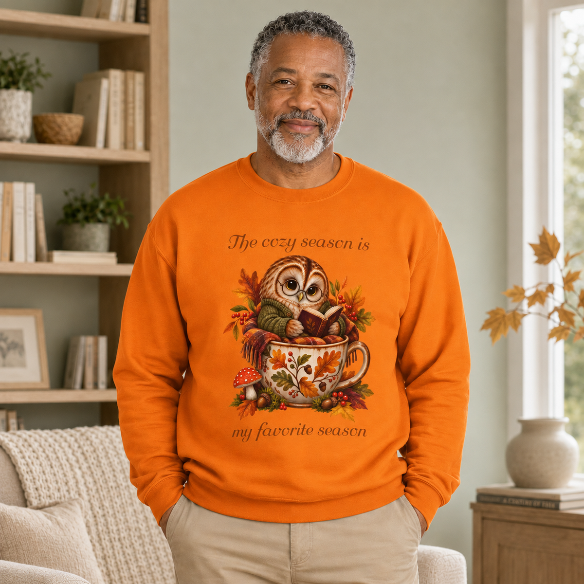 A man wearing a bright orange pullover with a graphic of an owl wearing a sweater and bundled in a blanket while reading a book in a teacup surrounded by autumn leaves and the text, 'The cozy season is my favorite season'.