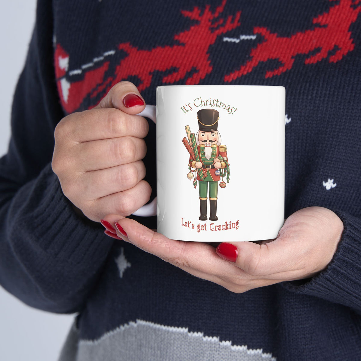 A person holding a white ceramic mug with a Christmas nutcracker illustration and the text 'It's Christmas' and 'Let's get Cracking'. The person is wearing a navy blue knit sweater with a red reindeer.