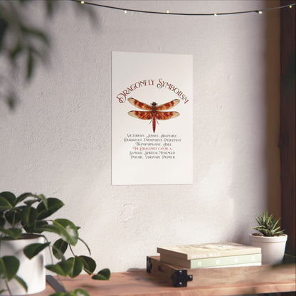 A vertical wall art print featuring a red dragonfly with the title 'Dragonfly Symbolism' and explanatory text below, displayed on a textured light wall above a wooden surface with books and plants.