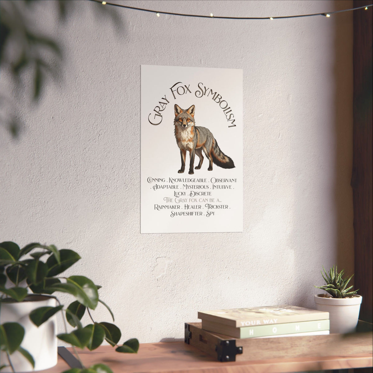 A vertical wall art print featuring a fox illustration and the text 'Gray Fox Symbolism' hanging on a textured wall above a wooden shelf with books and small plants.