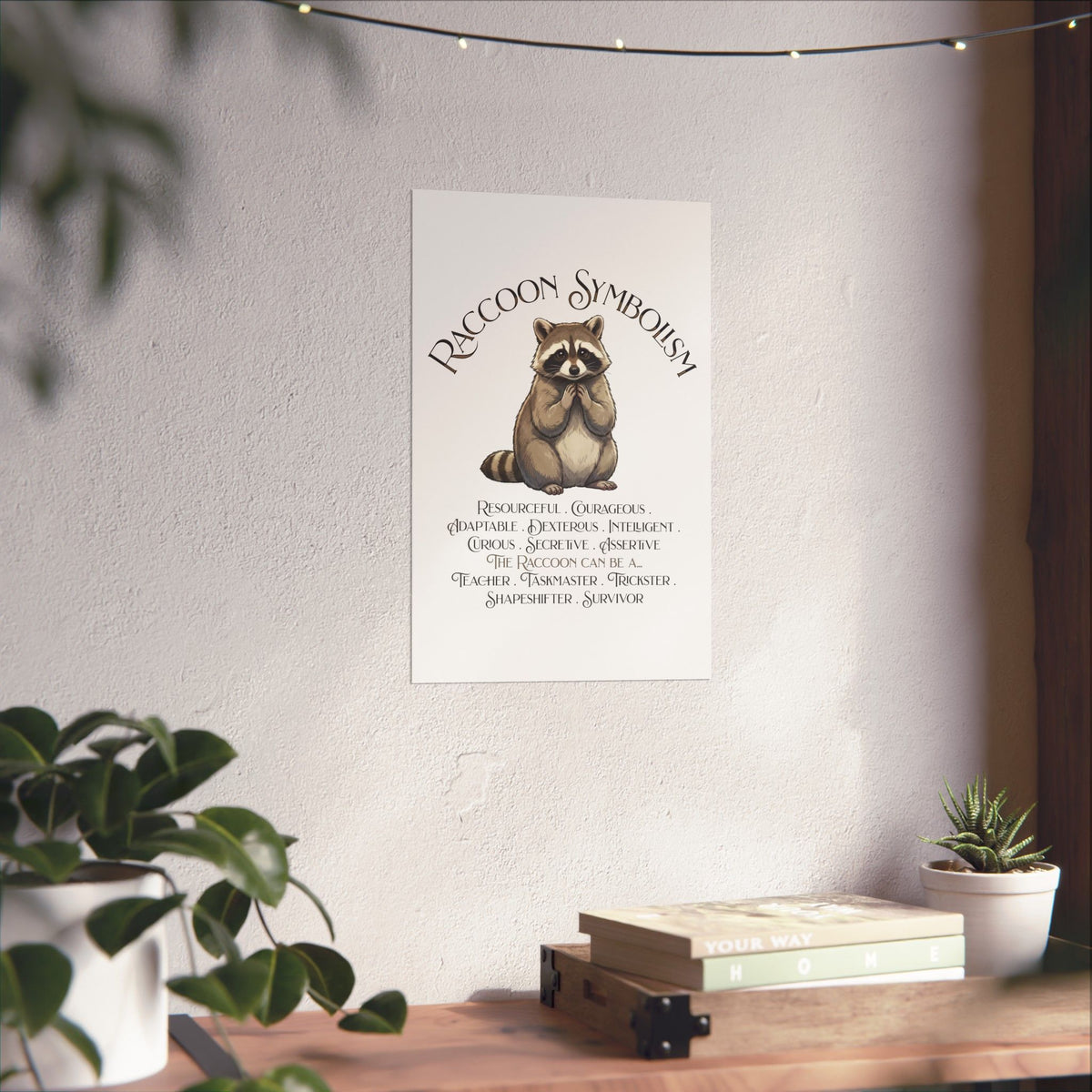 A vertical wall art print featuring a raccoon illustration and the text 'RACCOON SYMBOLISM' with traits listed below, displayed on a light wall above a wooden shelf with plants and books.