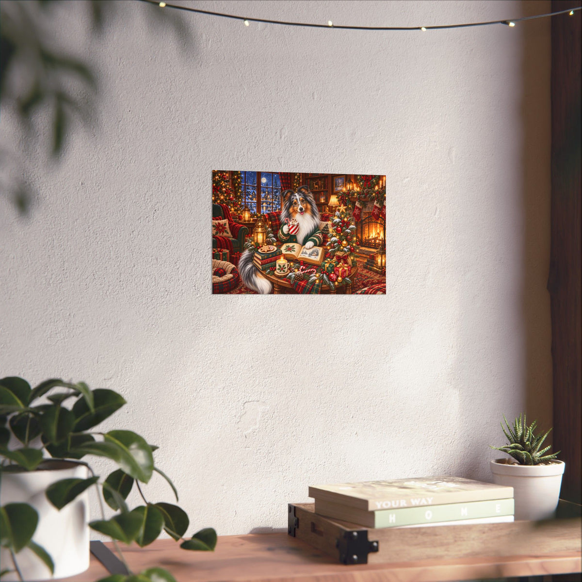 A rectangular wall art print showing a cozy Christmas scene with merle sheltie, displayed on a textured off-white wall above a wooden table with books and a small plant.