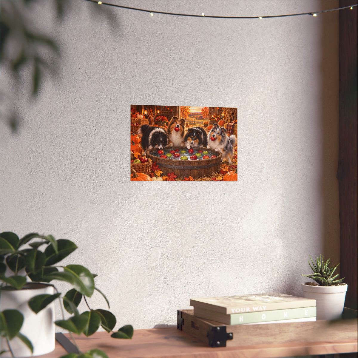 A rectangular wall art print showing four shelties bobbing for apples in an autumn harvest barn, displayed on a white wall above a shelf with books and potted plants.