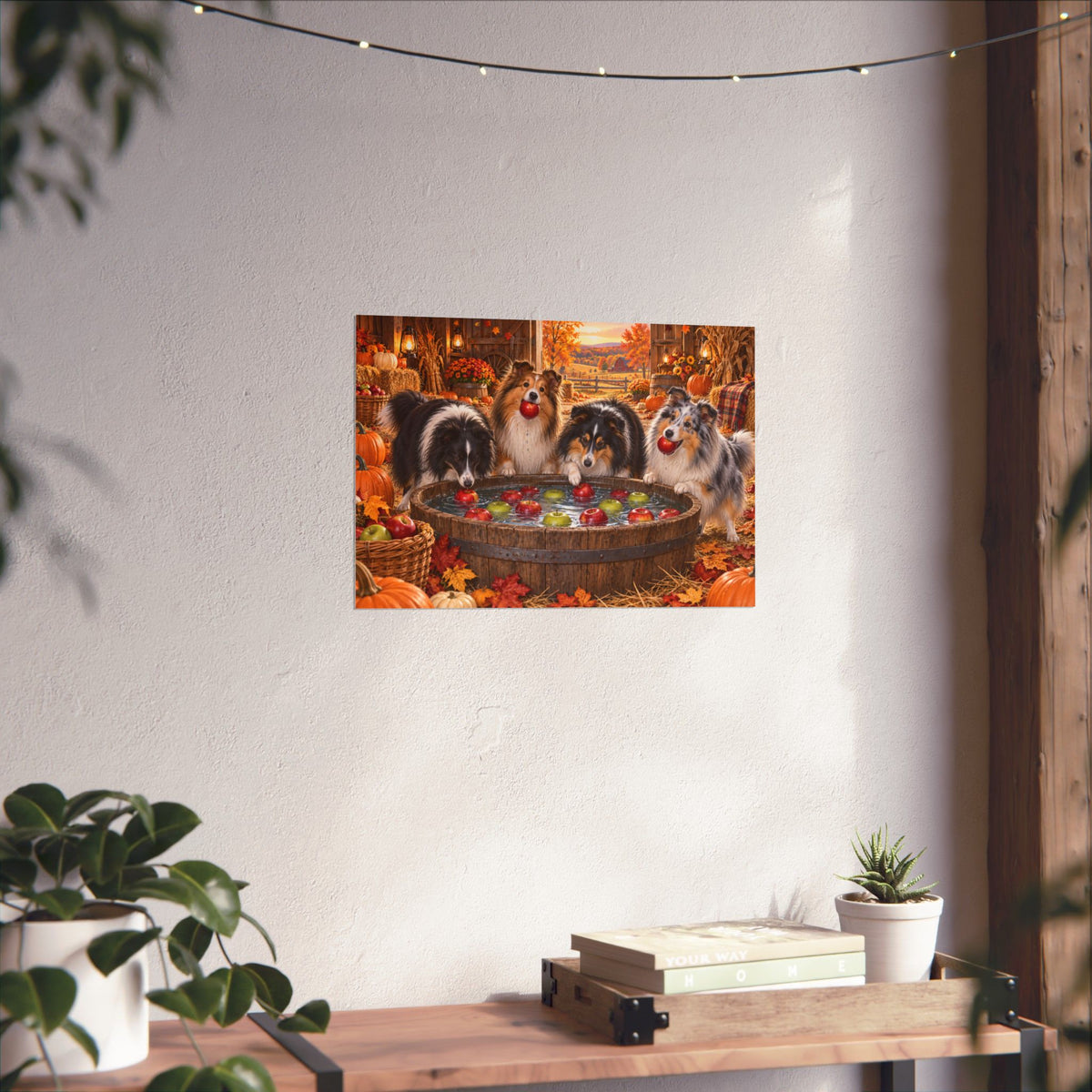 A rectangular wall art print showing four shelties bobbing for apples in an autumn harvest barn, displayed on a white wall above a shelf with books and potted plants.