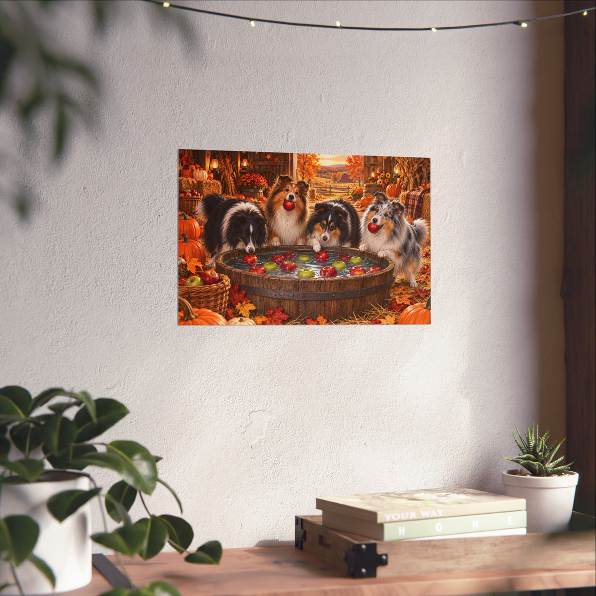 A rectangular wall art print showing four shelties bobbing for apples in an autumn harvest barn, displayed on a white wall above a shelf with books and potted plants.
