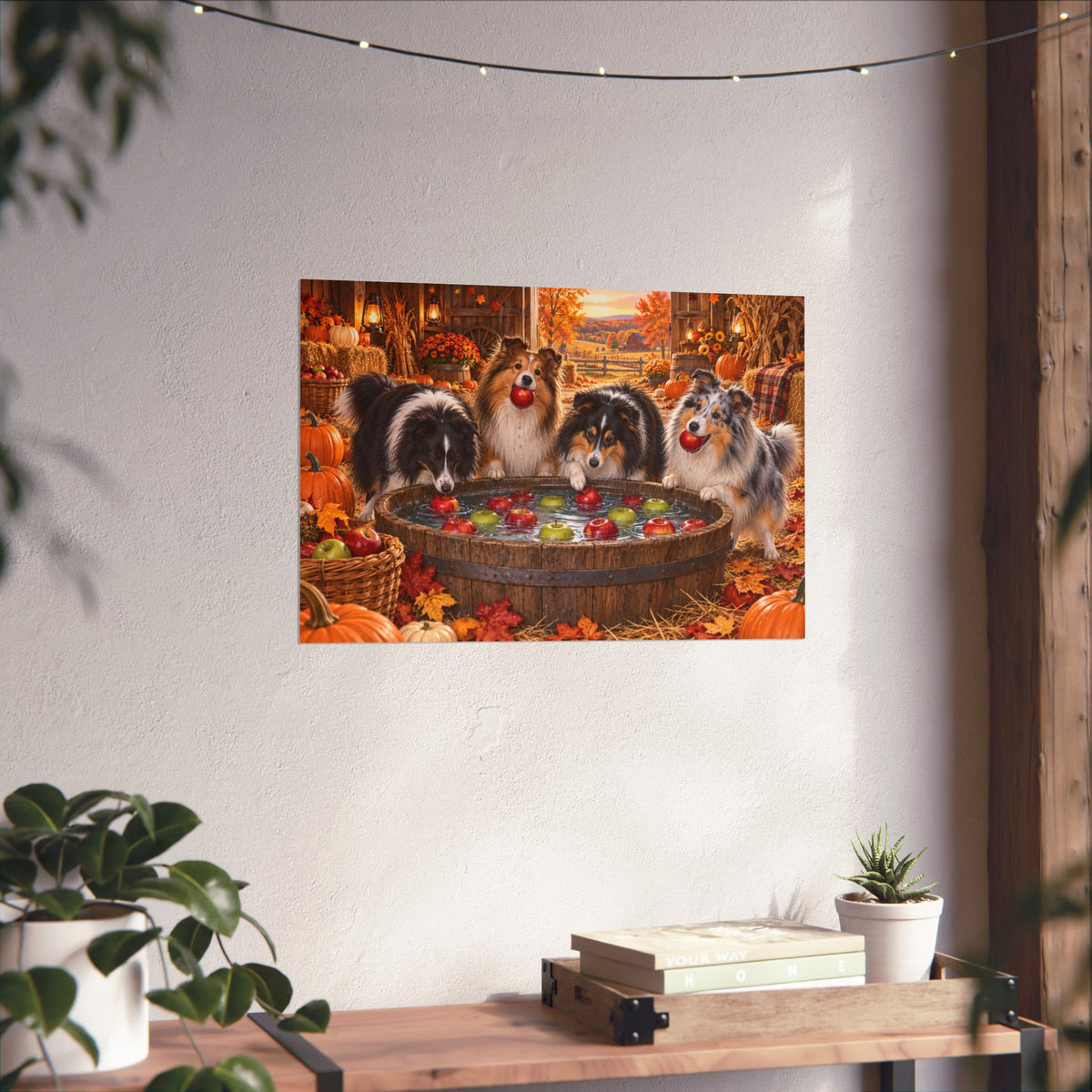 A rectangular wall art print showing four shelties bobbing for apples in an autumn harvest barn, displayed on a white wall above a shelf with books and potted plants.