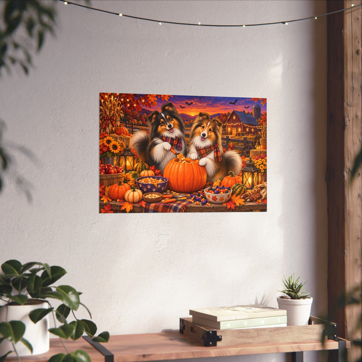 A rectangular wall art print showing two shelties carving a jack o' lantern, displayed on a white wall above a wooden surface with books and small plants.