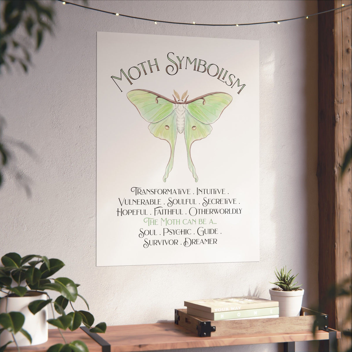 A vertical wall art print featuring a green luna moth illustration and the text 'Moth Symbolism' with meanings below, displayed on a textured wall in a styled room with plants and a wooden shelf.