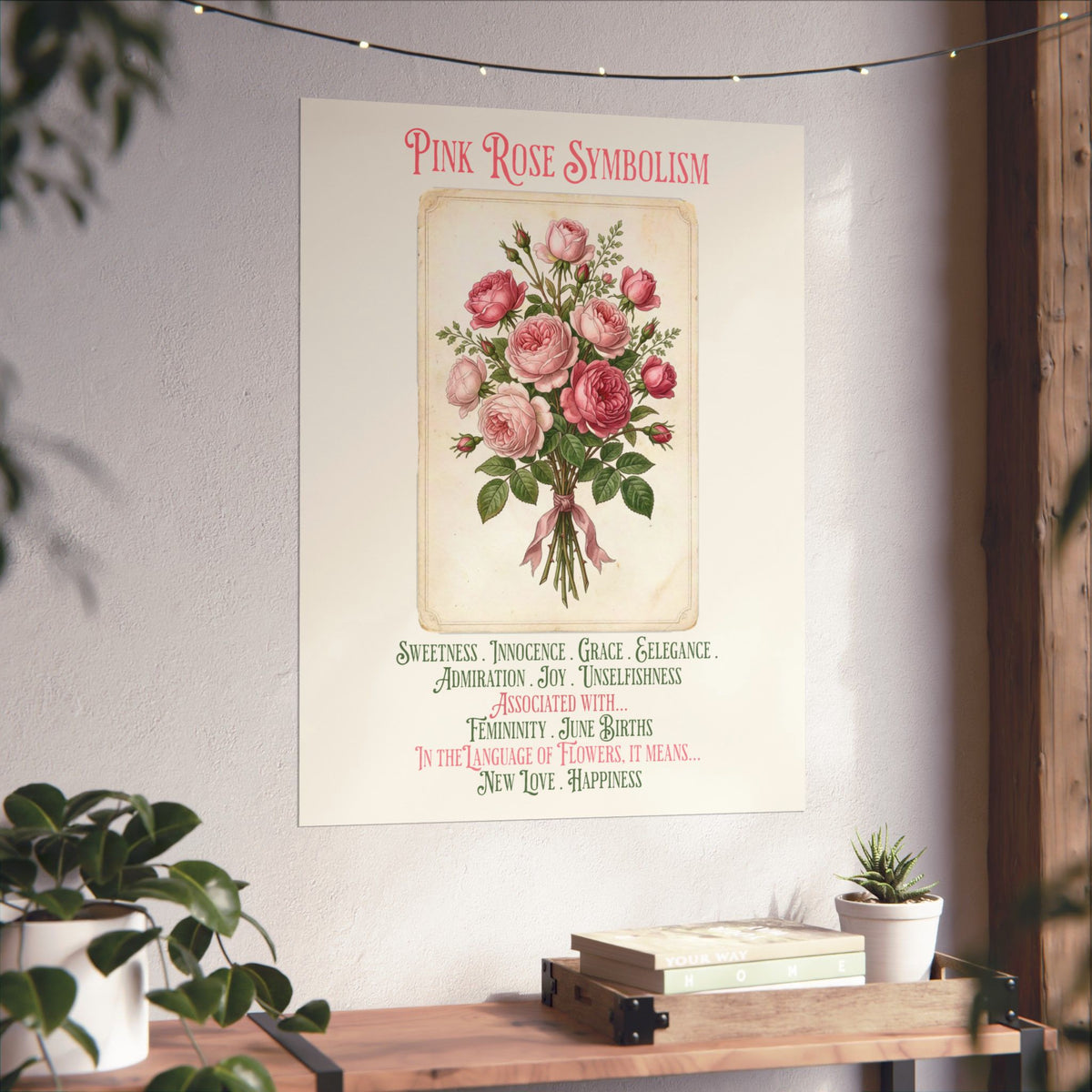 A vertical wall art print featuring a vintage-style illustration of pink roses with the title 'Pink Rose Symbolism' and decorative text below, displayed on a light wall above a wooden console with plants and books.