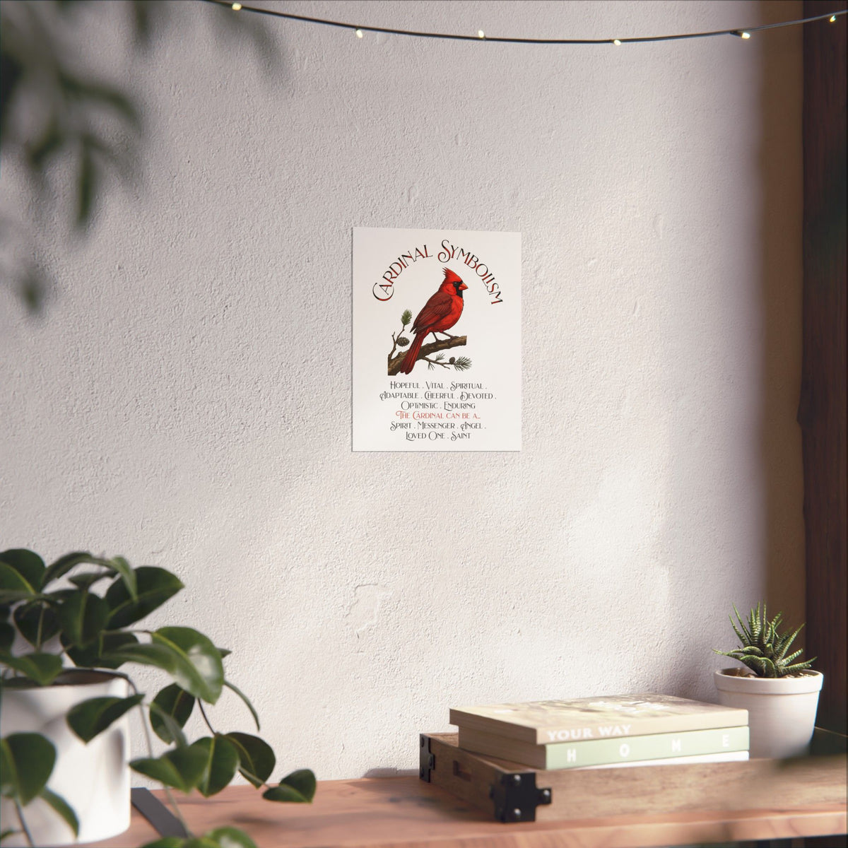 A vertical wall art print featuring a red cardinal on a pine branch with the title 'Cardinal Symbolism' and a list of cardinal qualities and meanings, displayed on a light wall above a wooden shelf with books and plants.