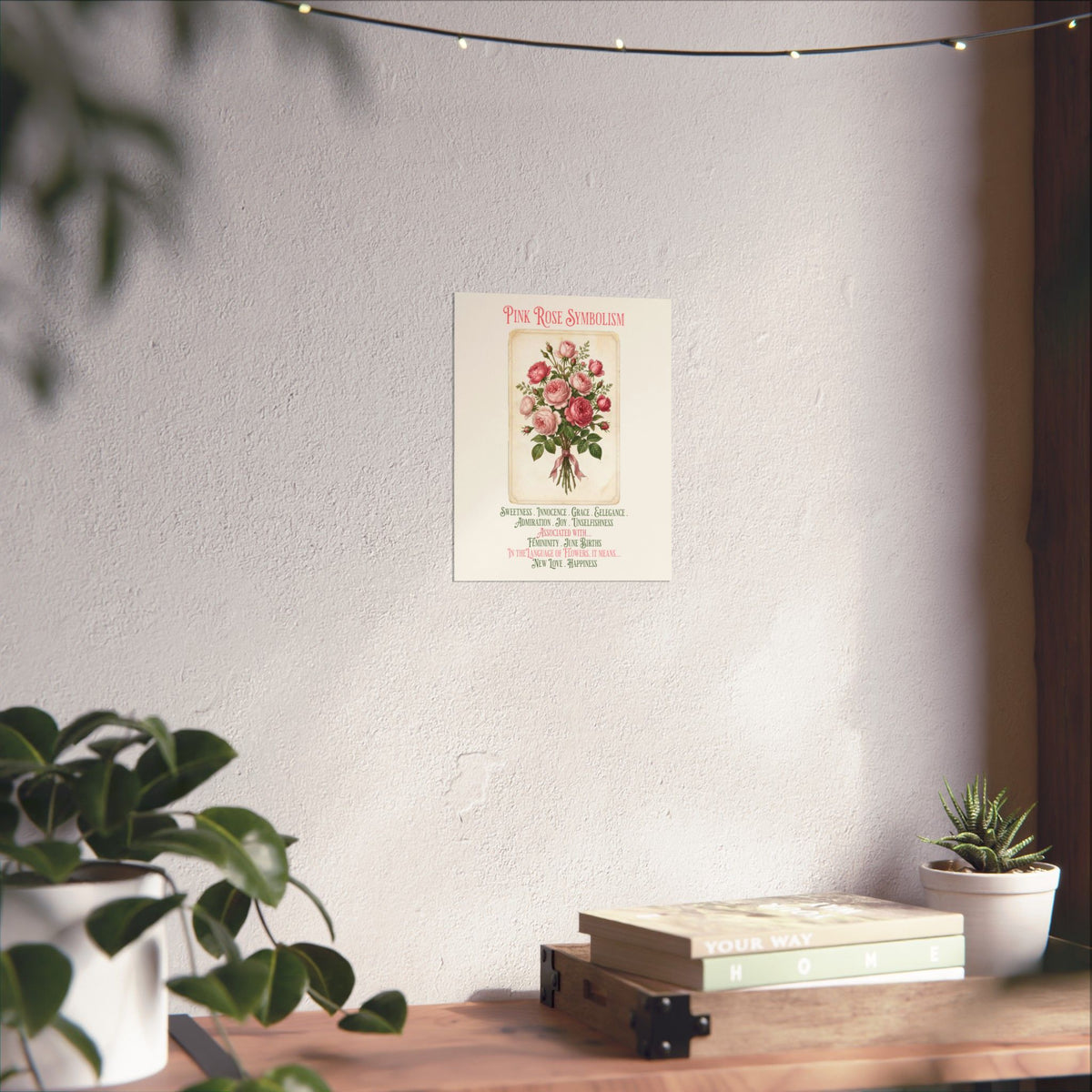 A vertical wall art print featuring a vintage-style illustration of pink roses with the title 'Pink Rose Symbolism' and decorative text below, displayed on a light wall above a wooden console with plants and books.
