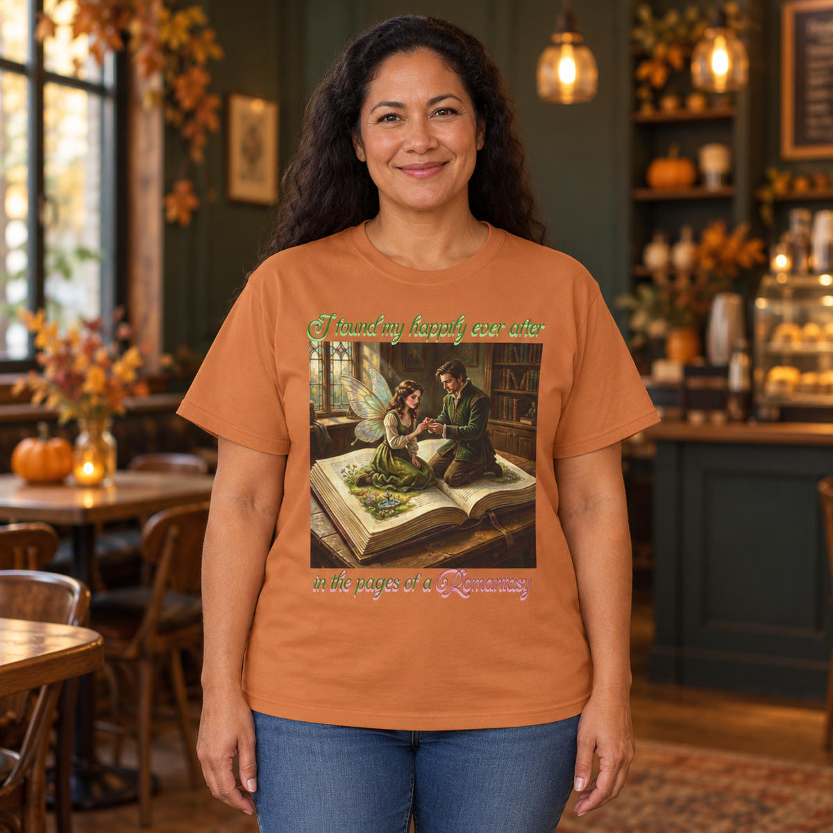 A woman wearing a burnt orange graphic t-shirt with a vintage proposal illustration and the text 'I found my happily ever after' above and below, 'in the pages of a Romantasy'. The woman is standing in a cozy, warmly lit cafe.