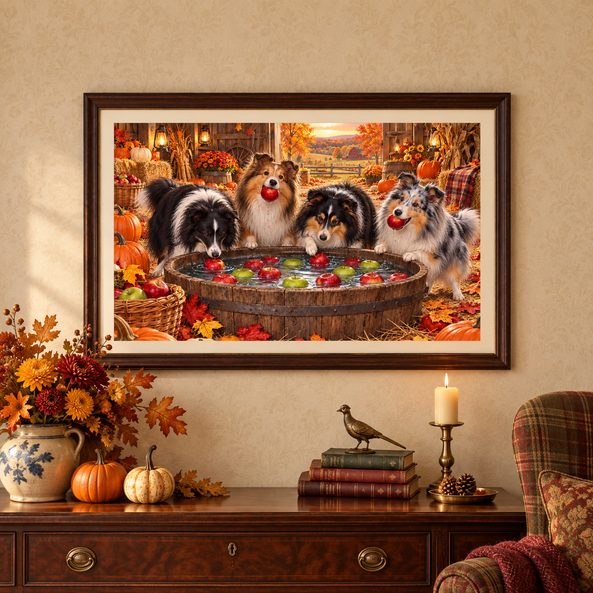 A framed wall art print depicting four shelties bobbing for apples in an autumn barn, displayed on a pale golden wallpaper above a mahogany console table with a vase of marigolds and fall leaves, stack of books, a brass bird figure, and a lit candle on a brass stand. A plaid chair with a burgundy blanket and a red and gold leaf patterned pillow sits nearby.