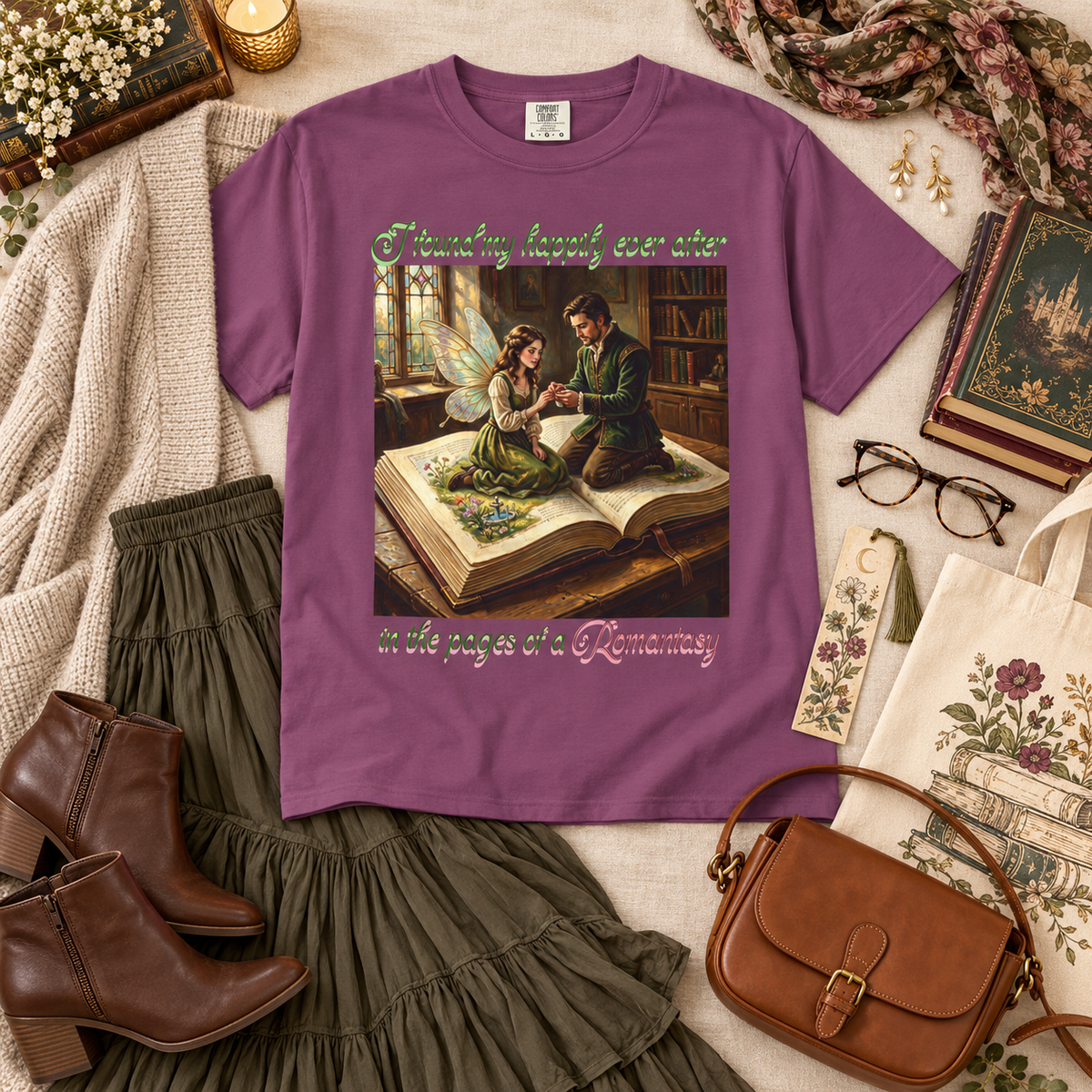 A purple graphic T-shirt laid flat on a styled surface with a cozy book-and-fairy illustration on the front. The shirt includes decorative text reading 'Finding happy ever after in the pages of a Romantasy'. The shirt is surrounded by books, a book bag, a bookmark, glasses, and a feminine outfit ensemble.