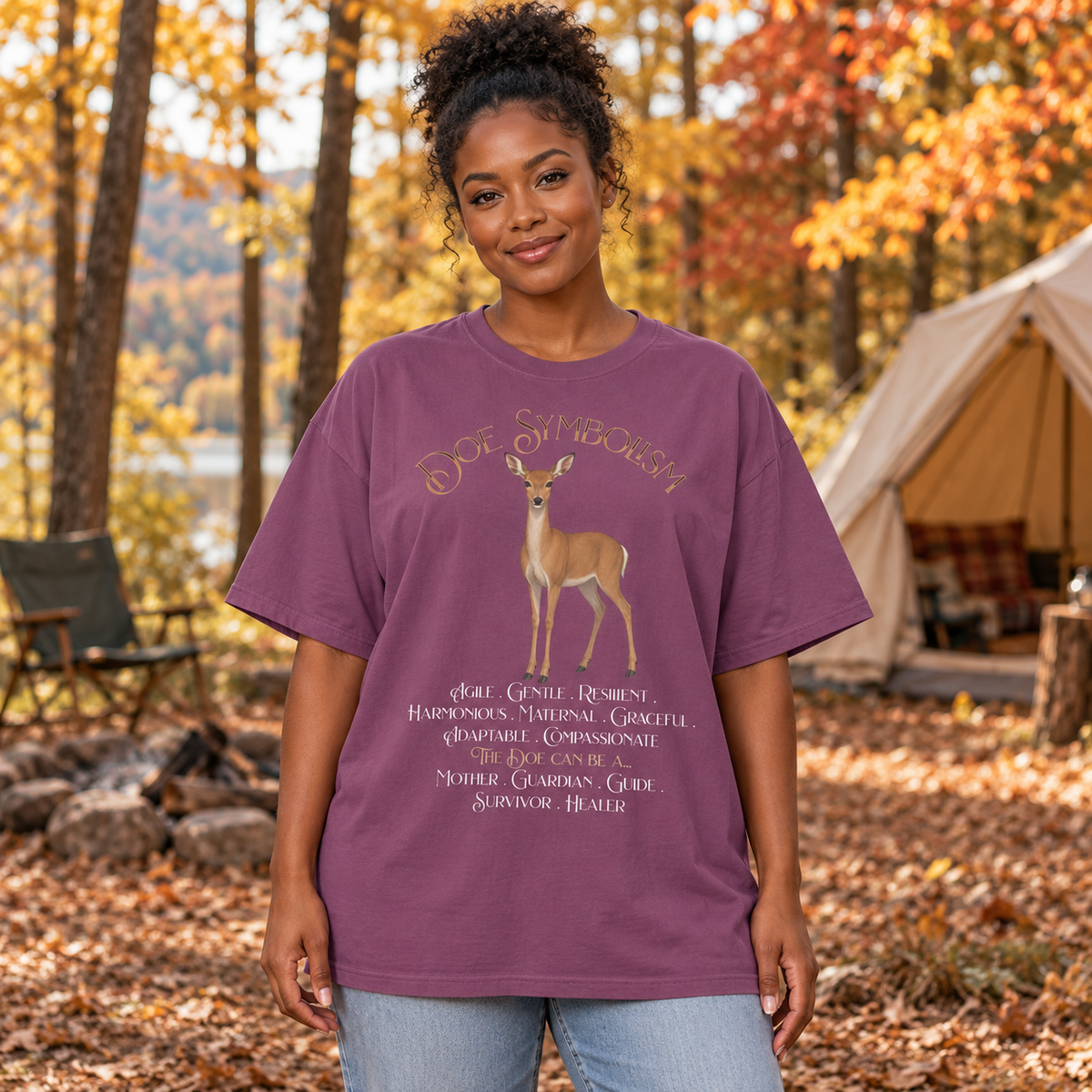A woman wearing an oversized purple graphic t-shirt with a deer illustration and the title Doe Symbolism followed by descriptive text. The woman stands in an autumn forest with a tent in the background.