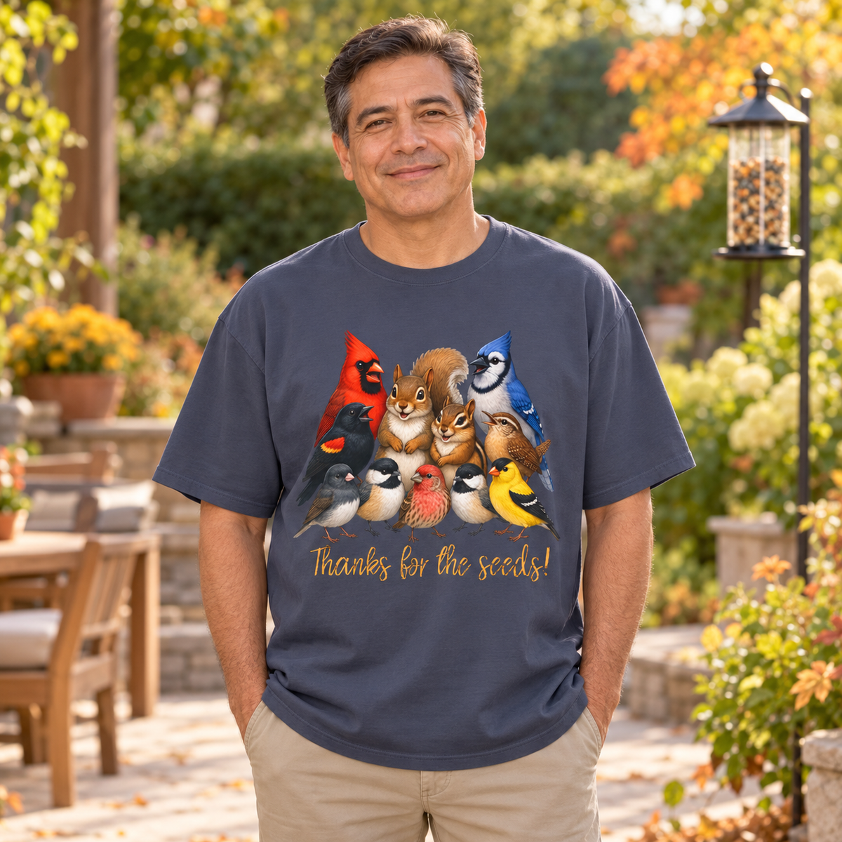 A man wearing a navy blue t-shirt with a colorful graphic of various birds, a squirrel, and a chipmunk above the text 'Thanks for the seeds!' The man is in a sunny outdoor garden setting.
