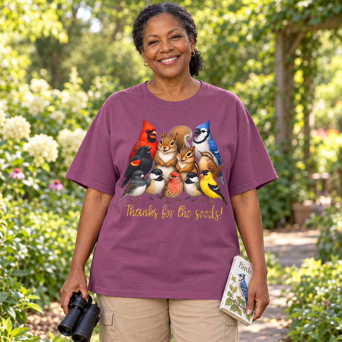 A smiling woman stands outdoors in a garden wearing a purple graphic T-shirt with a colorful group of birds, chipmunk, and a squirrel and the text 'Thanks for the seeds!' She is holding black binoculars in one hand and a small bird book in the other.