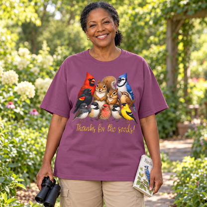 A smiling woman stands outdoors in a garden wearing a purple graphic T-shirt with a colorful group of birds, chipmunk, and a squirrel and the text 'Thanks for the seeds!' She is holding black binoculars in one hand and a small bird book in the other.