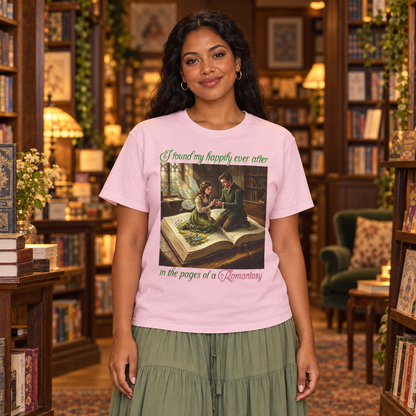 A woman wearing a light pink graphic t-shirt with a romantic fairy and man illustration and the text 'I found my happily ever after in the pages of a Romantasy'. The woman is standing in a cozy, vintage library setting.