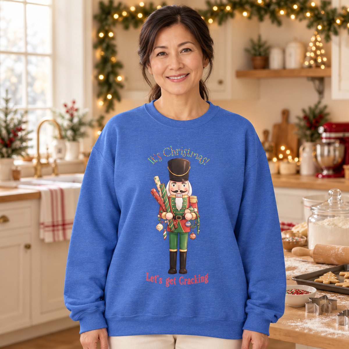 A woman wearing a royal blue crewneck sweatshirt with a festive nutcracker graphic and the text 'It's Christmas! Let's get Cracking' in a warm, decorated kitchen setting.
