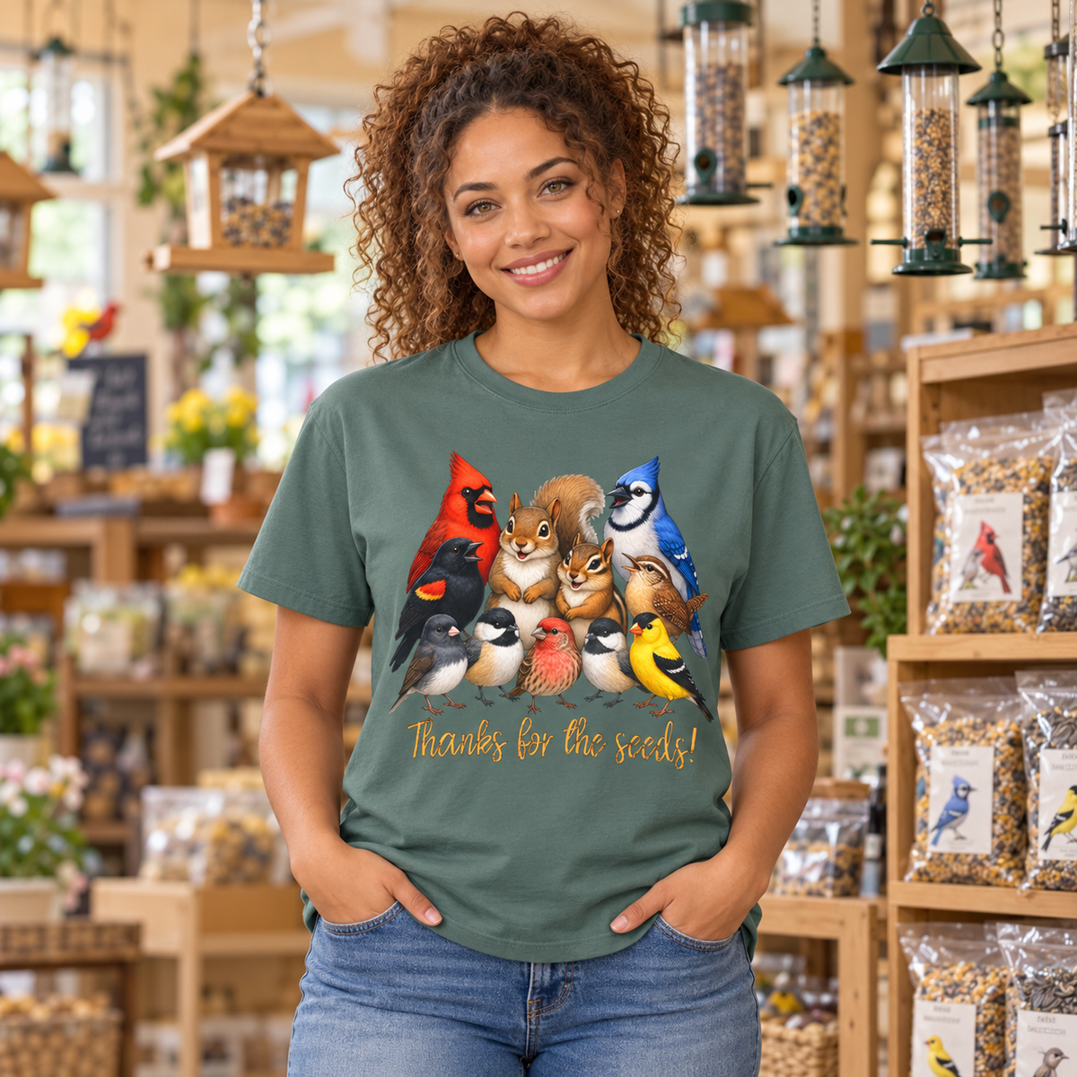 A woman with curly brown hair stands in a bird store wearing a sage green graphic t-shirt featuring a colorful illustration of various birds, a chipmunk, and a squirrel and the text 'Thanks for the seeds!'