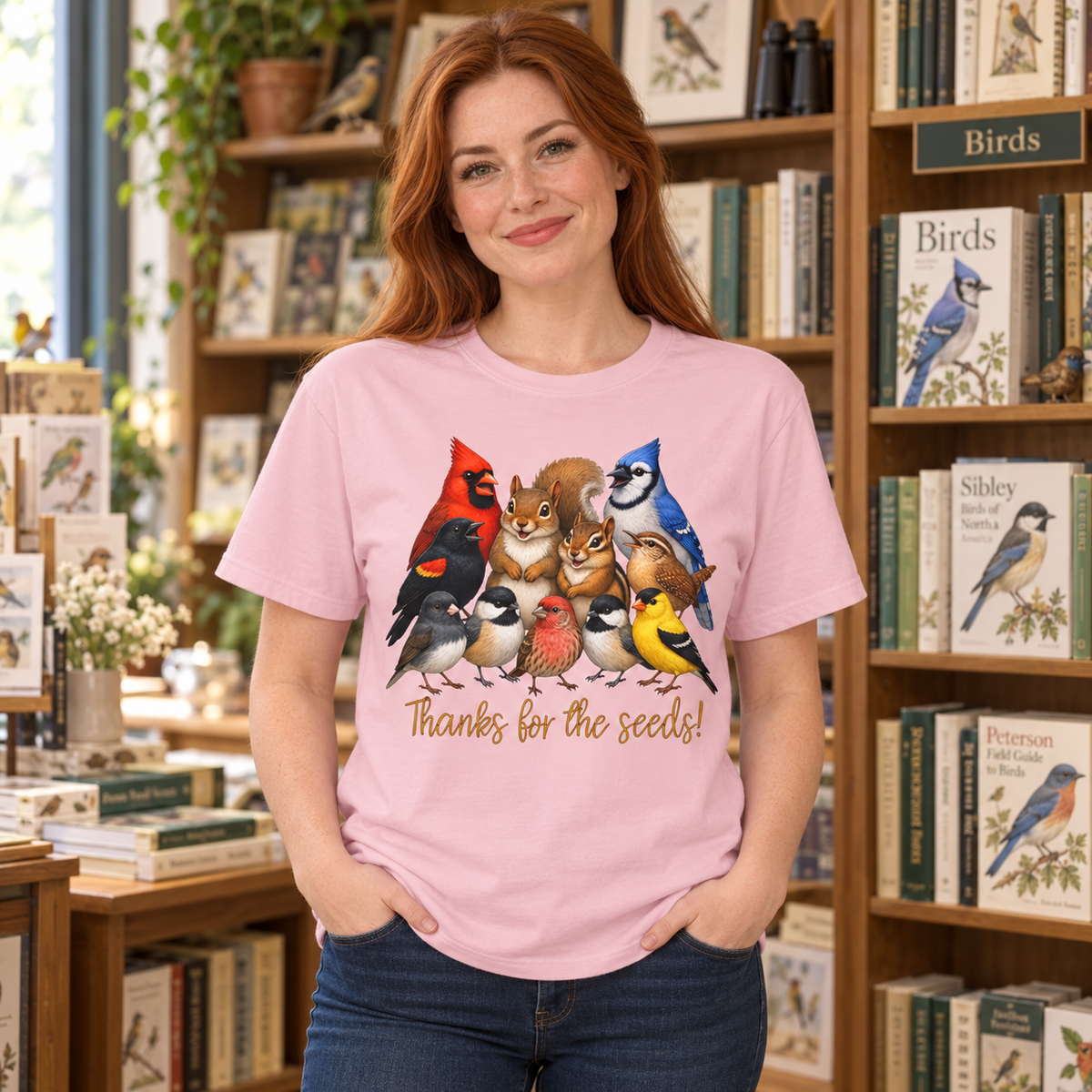 A woman with red hair stands in a bookshop wearing a light pink t-shirt with a colorful graphic of various birds, a chipmunk, and a squirrel  and the text 'Thanks for the seeds!'