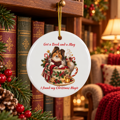 A round white ceramic Christmas ornament hanging from a gold string in a cozy holiday room. The ornament features an illustration of a Sheltie reading a book in a teacup, with red text reading 'Got a Book and a Mug I found my Christmas Magic'.