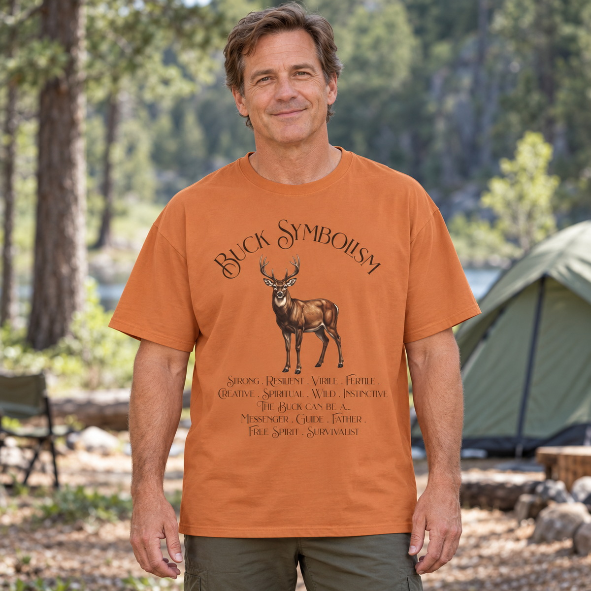 A man wearing an orange graphic t-shirt with a deer design and the text 'Buck Symbolism' and a list of its symbolic meanings in an outdoor forest setting with a tent in the background.